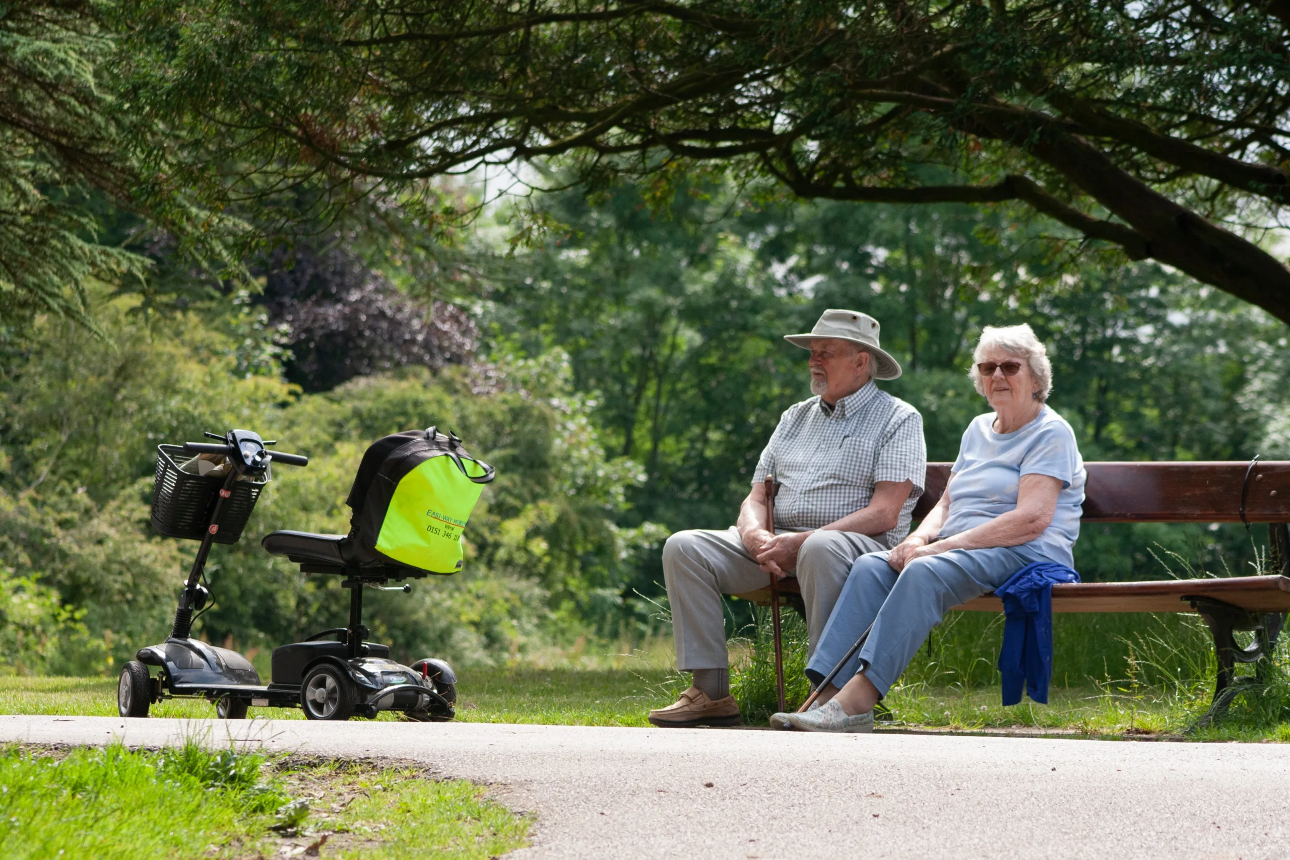 Mature Couple Sat On Park Bench Next To Mobility Scooter